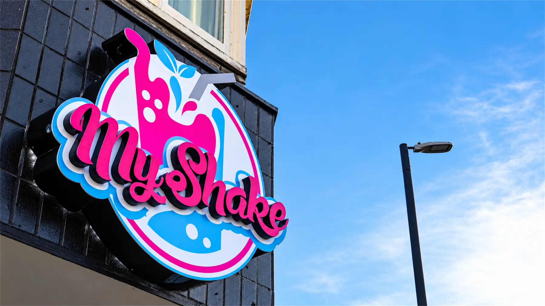Round 3D logo sign for a milkshake shop in pink, white and blue, mounted on a dark tiled wall